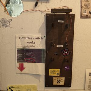 A double knife switch made of copper and a base of wood placed on a wall full of posters and stickers. It is on the outside of the printer room near the front entrance